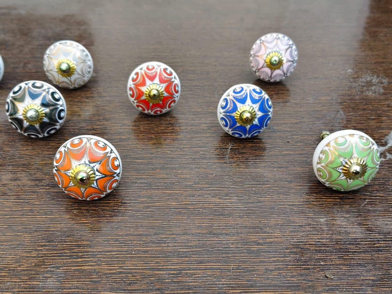 Pu&ograve; includere: Una collezione di sette pomelli rotondi in ceramica per mobili con intricati motivi floreali multicolori nelle tonalit&agrave; arancione, rosso, blu, verde, nero e bianco, ciascuno centrato con un piccolo dettaglio in metallo color oro.