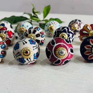 May include: A selection of decorative ceramic cabinet knobs with floral patterns. The knobs are in various colours, including red, blue, white, and gold. Each knob has a unique design and a gold-coloured metal base.