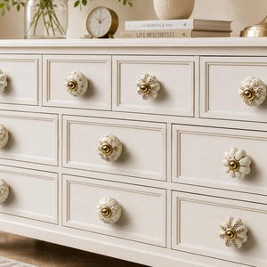 May include: A white wooden dresser featuring nine drawers, each equipped with a decorative ceramic knob. The knobs display various patterns in white and gold, including floral and geometric designs, with metallic gold centers and hardware.