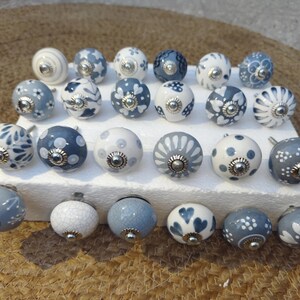 May include: A selection of decorative ceramic cabinet knobs in various designs. The knobs are mainly white and blue, featuring floral, polka dot, and heart patterns. Each knob has a silver-toned metal base and stem. The knobs are arranged on a white rectangular surface.