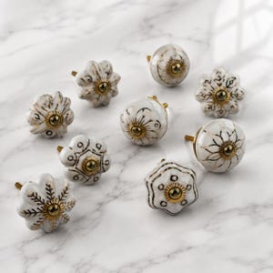 Pode incluir: Um conjunto de oito puxadores de cerâmica para armários com superfícies brancas e vários padrões florais ou geométricos pintados a preto e dourado. Cada puxador tem uma base de parafuso central em tom dourado e uma forma arredondada.