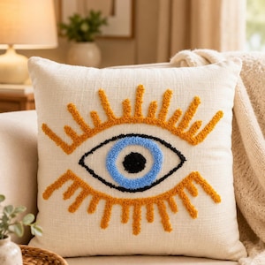 May include: Cream-colored decorative throw pillow featuring an embroidered eye design. The eye has a blue iris, black pupil, and gold eyelashes. The pillow is on a beige sofa, with a lamp and artwork in the background.
