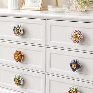 Op de afbeelding: Een witte ladekast met zes lades voorzien van decoratieve keramische knoppen. Elke knop heeft een uniek bloemmotief of geometrisch patroon in levendige kleuren zoals blauw, rood, geel en groen, met een gouden accent in het midden.