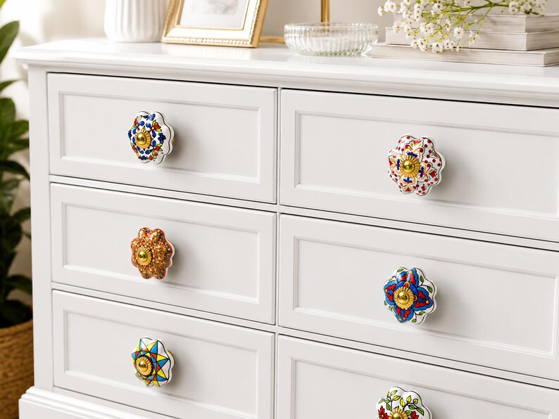 Op de afbeelding: Een witte ladekast met zes lades voorzien van decoratieve keramische knoppen. Elke knop heeft een uniek bloemmotief of geometrisch patroon in levendige kleuren zoals blauw, rood, geel en groen, met een gouden accent in het midden.