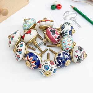 Pu&ograve; includere: Una collezione di pomelli decorativi in ceramica con ferramenta in ottone. I pomelli presentano vari disegni floreali e geometrici dipinti a mano in blu, rosso e bianco. Una matita, uno schizzo e una scatola di legno sono sullo sfondo.