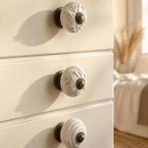 May include: Three round ceramic drawer knobs attached to a cream-coloured cabinet. Each knob features a white surface with embossed patterns and a dark bronze-toned metal base and centre cap. The top two knobs have floral motifs, while the bottom knob has concentric circular ridges.