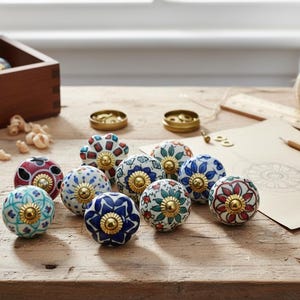 Pu&ograve; includere: Una collezione di pomelli decorativi in ceramica con disegni floreali in vari colori, tra cui blu, rosso e verde. Ogni pomello ha una base color oro. I pomelli sono disposti su una superficie di legno.