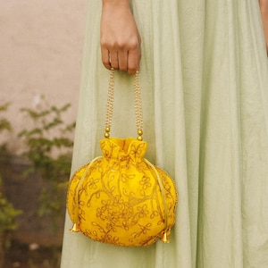 May include: A yellow drawstring bag with gold floral embroidery and beaded handles. The bag has a rounded shape and small gold tassels at the bottom. The bag is held by a person wearing a light green dress.