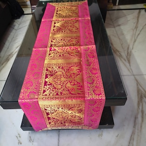 Könnte beinhalten: Dekorativer Tischläufer in leuchtendem Pink und Gold. Der Läufer zeigt ein kompliziertes Goldmuster auf tiefrosa Grund, mit einem kontrastierenden rosafarbenen Rand. Das Design umfasst Blumen- und Vogelmotive und verleiht jeder Tischdekoration Eleganz.
