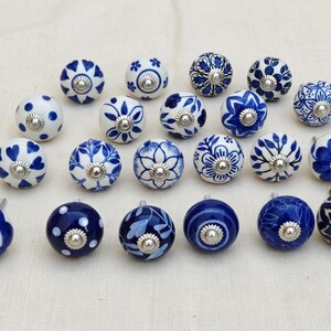 Può includere: Una collezione di venti pomelli rotondi in ceramica per mobili con vari motivi blu e bianchi, inclusi disegni floreali, geometrici e a pois. Ogni pomello è rifinito con un elemento metallico centrale per il fissaggio.
