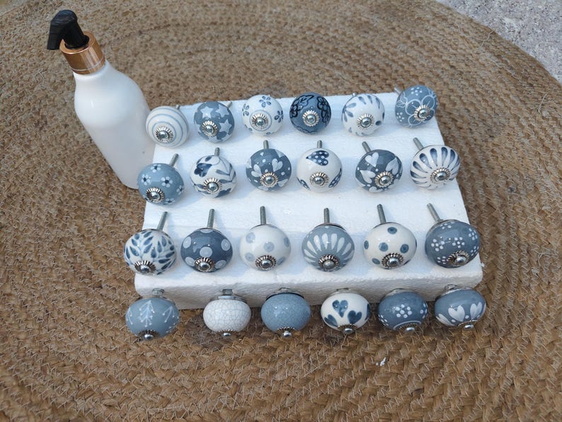 Op de afbeelding: Een verzameling decoratieve keramische kastknoppen in verschillende ontwerpen en kleuren, waaronder wit, blauw en grijs. De knoppen hebben bloemen-, stippen- en patroonontwerpen. Er is ook een witte fles met een zwarte en gouden dop aanwezig.