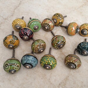 May include: A collection of fourteen round ceramic cabinet knobs featuring various hand-painted patterns in shades of green, yellow, black, and orange. Each knob has a metallic centre and a short metal bolt extending from the back for installation.
