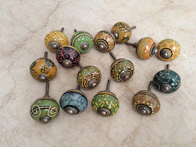 May include: A collection of fourteen round ceramic cabinet knobs featuring various hand-painted patterns in shades of green, yellow, black, and orange. Each knob has a metallic centre and a short metal bolt extending from the back for installation.