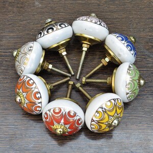 Pu&ograve; includere: Una collezione di sette pomelli rotondi in ceramica per mobili disposti in cerchio. Ogni pomello presenta una base bianca con distinti motivi colorati in arancione, giallo, rosso, verde, blu e grigio, accentuati da finiture in metallo dorato.