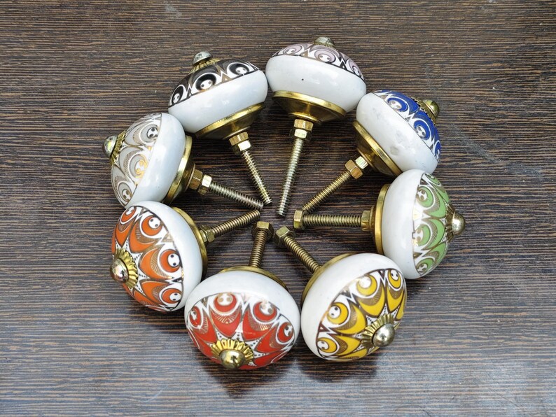 Pu&ograve; includere: Una collezione di sette pomelli rotondi in ceramica per mobili disposti in cerchio. Ogni pomello presenta una base bianca con distinti motivi colorati in arancione, giallo, rosso, verde, blu e grigio, accentuati da finiture in metallo dorato.