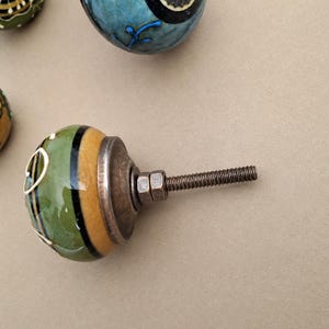 May include: A round ceramic cabinet knob featuring a glossy finish with alternating bands of olive green and mustard yellow, accented by thin black stripes and white line details. It includes a metallic base with a threaded bolt and two nuts.
