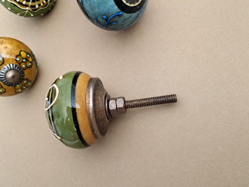 May include: A round ceramic cabinet knob featuring a glossy finish with alternating bands of olive green and mustard yellow, accented by thin black stripes and white line details. It includes a metallic base with a threaded bolt and two nuts.
