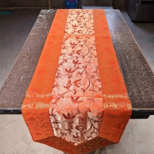 Op de afbeelding: Oranje tafelloper met een centraal paneel met een bloemmotief in rood en goud. De randen van de loper hebben een gedetailleerd goudkleurig patroon, wat een vleugje elegantie toevoegt aan het woondecoratie-item.