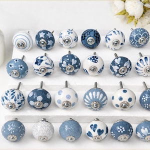 Può includere: Una collezione di pomelli decorativi in ceramica con vari motivi blu e bianchi. I pomelli rotondi hanno finiture argentate e sono disposti su un espositore bianco. A sinistra è visibile un dispenser di sapone dorato.