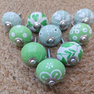 Pode incluir: Uma cole&ccedil;&atilde;o de dez puxadores decorativos de cer&acirc;mica em tons de verde e branco. Cada puxador apresenta um design &uacute;nico pintado &agrave; m&atilde;o, incluindo bolinhas, cora&ccedil;&otilde;es e padr&otilde;es florais. Os puxadores t&ecirc;m bases de metal prateado e est&atilde;o dispostos.
