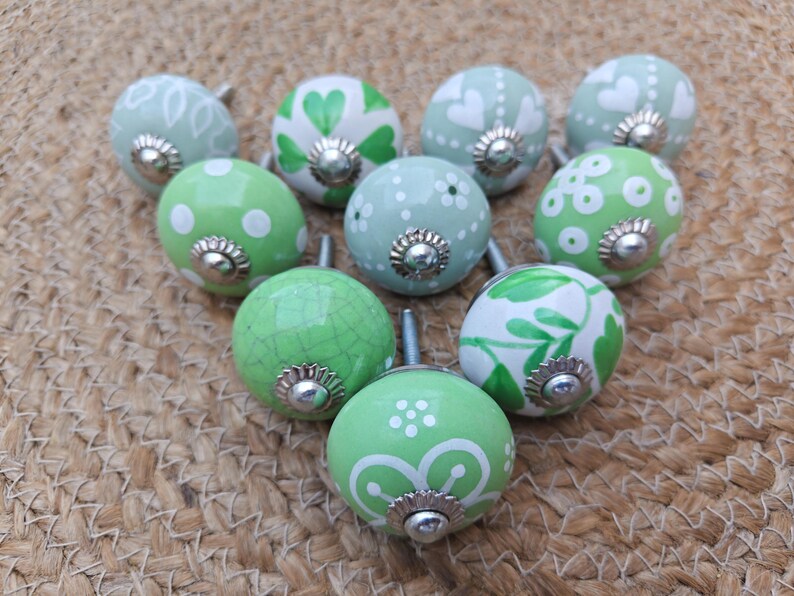 Pode incluir: Uma cole&ccedil;&atilde;o de dez puxadores decorativos de cer&acirc;mica em tons de verde e branco. Cada puxador apresenta um design &uacute;nico pintado &agrave; m&atilde;o, incluindo bolinhas, cora&ccedil;&otilde;es e padr&otilde;es florais. Os puxadores t&ecirc;m bases de metal prateado e est&atilde;o dispostos.