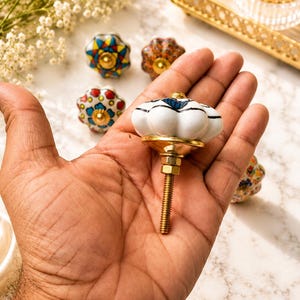 Op de afbeelding: Een witte keramische meubelknop met een geschulpte, pompoenachtige vorm en blauwe accenten. De knop heeft een goudkleurige metalen voet en een draadeind met een moer. Diverse andere kleurrijke, gedessineerde keramische knoppen zijn zichtbaar.