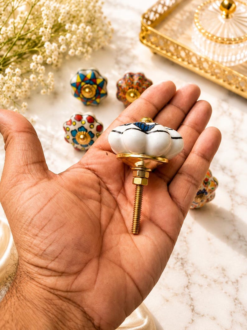 Op de afbeelding: Een witte keramische meubelknop met een geschulpte, pompoenachtige vorm en blauwe accenten. De knop heeft een goudkleurige metalen voet en een draadeind met een moer. Diverse andere kleurrijke, gedessineerde keramische knoppen zijn zichtbaar.