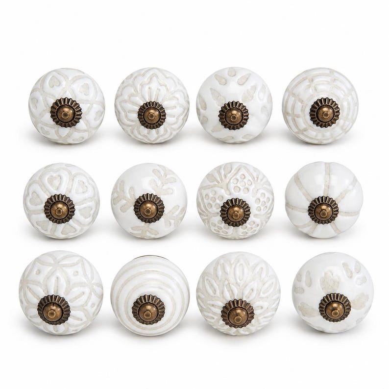 May include: A set of twelve round ceramic cabinet knobs arranged in three rows of four. Each knob features a white base with light grey embossed patterns and a central antique brass-coloured metal hardware piece with a fluted circular base.