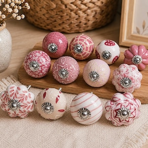 May include: A collection of twelve decorative cabinet knobs in various shades of pink and white. The knobs feature diverse patterns including floral motifs, geometric lines, and solid finishes, each with a central silver-toned metal hardware piece.