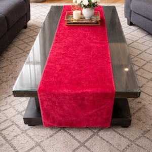 May include: A red velvet table runner is draped across a dark wood coffee table. A gold tray with a candle and a vase of white flowers sits on the runner. The table is in a living room with gray sofas and a patterned rug.