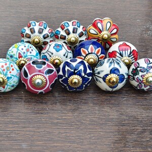 Pu&ograve; includere: Una collezione di undici pomelli decorativi in ceramica per armadi. I pomelli presentano vari motivi floreali e geometrici in colori come rosso, blu, bianco e verde. Ogni pomello ha una base in metallo dorato.