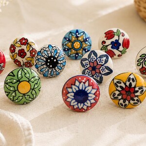 May include: A collection of twelve round ceramic cabinet knobs, each featuring unique hand-painted floral and geometric patterns in vibrant colors including red, blue, green, and yellow, arranged in a group on a light-colored fabric surface.