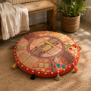 May include: A round floor cushion featuring a patchwork design of orange, gold, and multi-colored fabric panels with intricate embroidery, floral patterns, and metallic thread accents. The edge is decorated with small white shells and tassels.