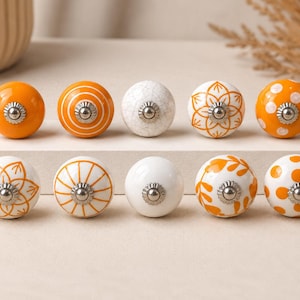 Op de afbeelding: Een set van tien ronde keramische kastknoppen gerangschikt in twee rijen. De knoppen hebben verschillende ontwerpen, waaronder effen oranje, wit met oranje stippen, oranje met witte strepen, wit met oranje bloemmotieven en een witte craquelé afwerking.