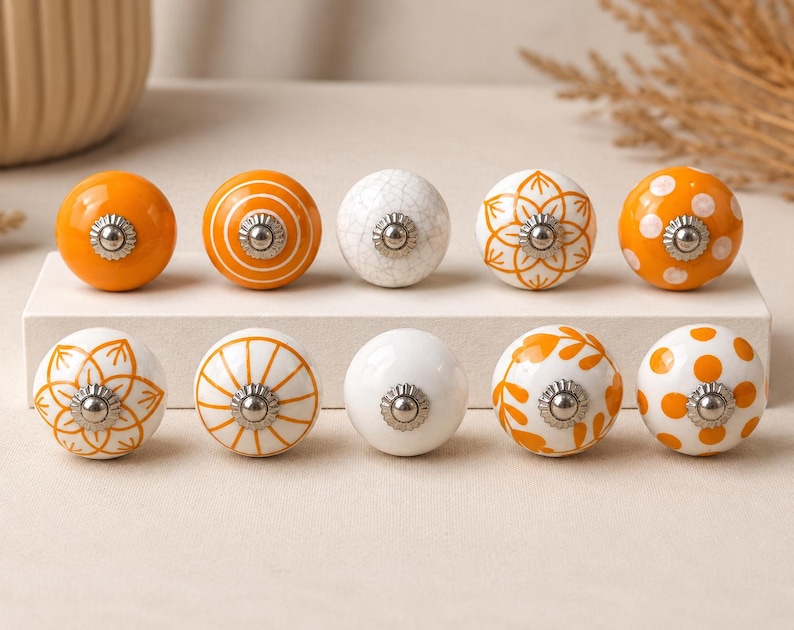 May include: A set of ten round ceramic cabinet knobs arranged in two rows. The knobs feature various designs including solid orange, white with orange polka dots, orange with white stripes, white with orange floral patterns, and a white crackle finish.