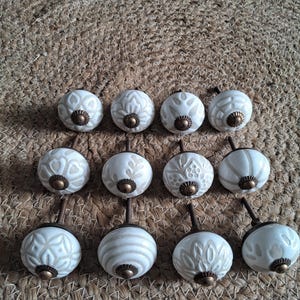 May include: A collection of twelve round ceramic cabinet knobs arranged in three rows. Each knob features a white base with light beige embossed patterns and a central antique brass-toned metal hardware piece with a threaded screw post.
