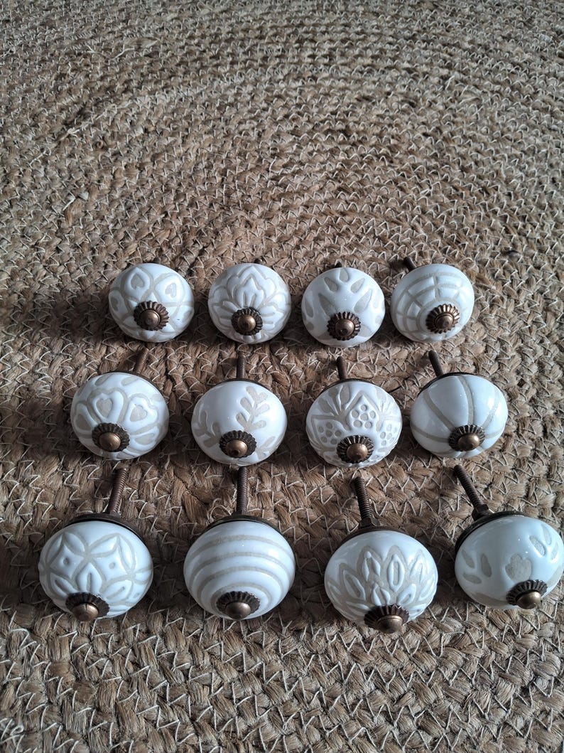 May include: A collection of twelve round ceramic cabinet knobs arranged in three rows. Each knob features a white base with light beige embossed patterns and a central antique brass-toned metal hardware piece with a threaded screw post.
