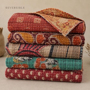 Op de afbeelding: Een stapel van vijf gevouwen omkeerbare quilts met fijn kantha-stiksel. De patronen bevatten rode geometrische vierkanten, oranje paisley, gestreepte texturen, groenblauwe bloemmotieven en kleine crèmekleurige paisleyprints op een rode achtergrond.