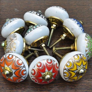 Pu&ograve; includere: Una collezione di dieci pomelli rotondi in ceramica per mobili con vari motivi geometrici colorati in arancione, rosso, giallo, verde e blu. Ogni pomello ha una base bianca, un centro in metallo dorato e un bullone filettato per il fissaggio.