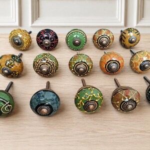May include: A collection of fourteen round ceramic cabinet knobs featuring various hand-painted patterns in shades of green, yellow, orange, and black. Each knob has a central metallic hardware piece with a screw stem for mounting purposes.