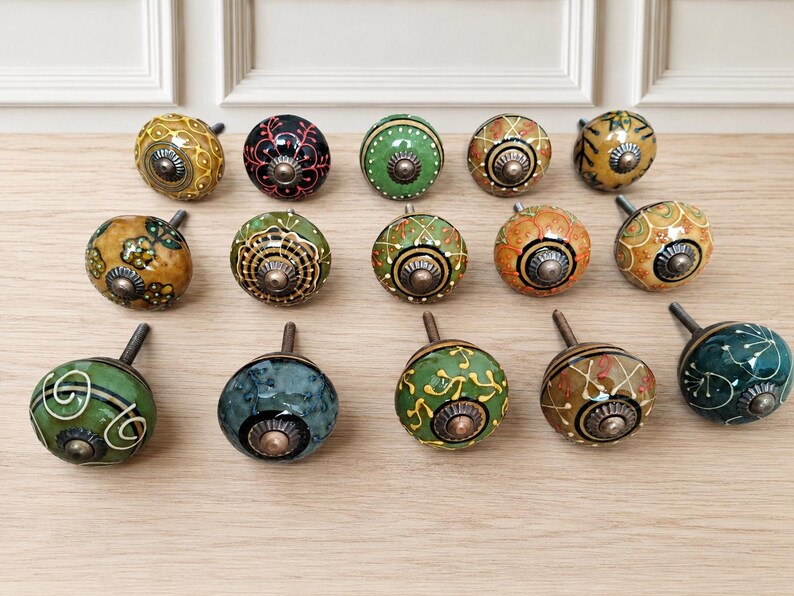 May include: A collection of fourteen round ceramic cabinet knobs featuring various hand-painted patterns in shades of green, yellow, orange, and black. Each knob has a central metallic hardware piece with a screw stem for mounting purposes.
