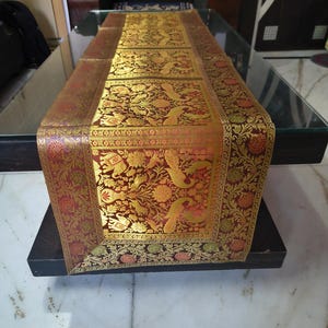 Könnte beinhalten: Dekorativer Tischläufer mit einem reichen, verzierten Design. Der Läufer hat einen tiefroten Hintergrund mit goldenen Blumen- und Vogelmotiven. Die Ränder sind mit einem goldenen und roten Muster eingefasst, das der Tischdekoration einen Hauch von Eleganz verleiht. Ca. 150 cm lang.