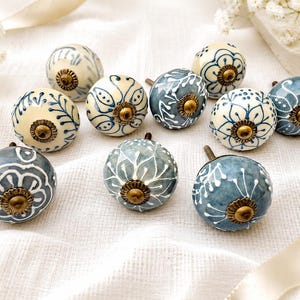 Può includere: Una collezione di dieci pomelli rotondi in ceramica per mobili con vari motivi floreali e botanici dipinti a mano in blu e bianco. Ogni pomello presenta un elemento centrale in metallo color ottone antico con una piccola vite sul retro.