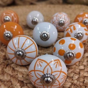 Pode incluir: Uma coleção de puxadores de armário decorativos em cerâmica. Os puxadores são brancos e laranja, apresentando vários padrões como bolinhas, listras e desenhos florais. Cada puxador tem uma base de metal prateado. Os puxadores estão dispostos sobre uma superfície tecida e texturizada.