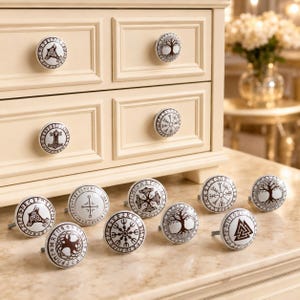 May include: A collection of round white ceramic drawer knobs featuring dark brown printed designs including Norse symbols, the Tree of Life, and runic patterns. Two knobs are installed on a cream-colored wooden chest, while nine others sit on a surface.