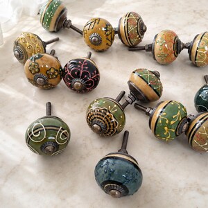 May include: A collection of twelve round ceramic cabinet knobs featuring various hand-painted designs in colours including green, yellow, black, and orange. Each knob has a metallic bolt and a decorative metal base with intricate patterns.