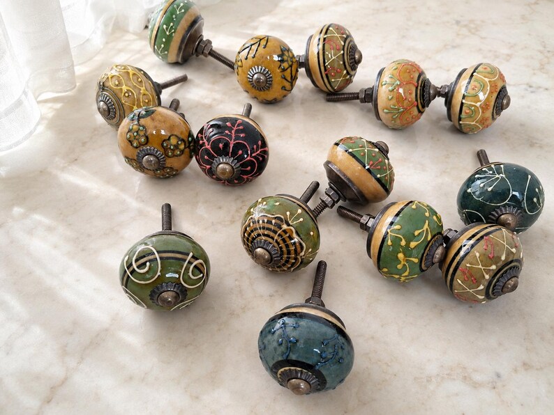 May include: A collection of twelve round ceramic cabinet knobs featuring various hand-painted designs in colours including green, yellow, black, and orange. Each knob has a metallic bolt and a decorative metal base with intricate patterns.