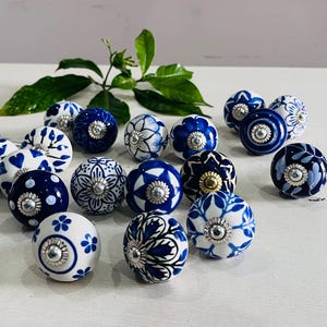 May include: A collection of decorative ceramic cabinet knobs in various designs. The knobs are primarily white and navy blue, featuring floral, polka dot, and heart patterns. Each knob has a silver metal base. A sprig of green leaves is in the background.