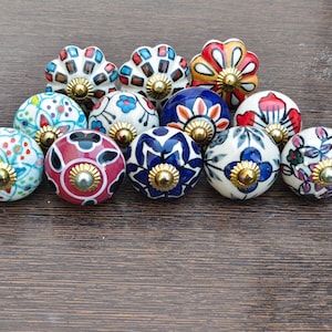 Può includere: Una collezione di pomelli decorativi in ceramica con motivi floreali e geometrici in vari colori, tra cui rosso, blu, bianco e verde. Ogni pomello ha una base metallica dorata.