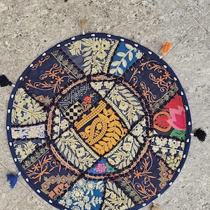 Könnte beinhalten: Ein rundes Bodenkissen mit einem Patchwork-Design aus verschiedenen dunkelblauen, goldenen und bunten Stoffbahnen mit aufwendigen Stickereien und metallischen Fadenakzenten. Der Rand ist mit einer dunkelblauen Borte und kleinen Quasten versehen.